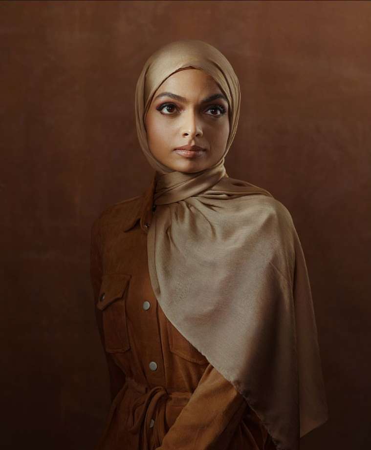 Portrait of a woman with dark hair wearing a brown hijab and matching jacket against a blended brown backdrop in a photo studio.